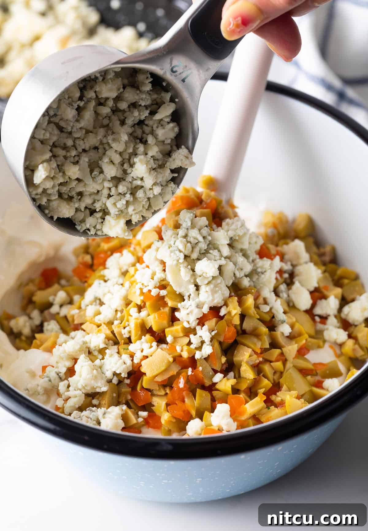 Adding crumbled blue cheese to a bowl containing chopped olives, sour cream, and cream cheese mixture.
