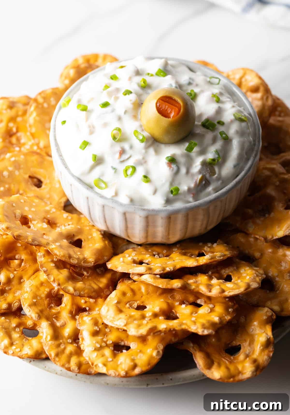 White bowl of dirty martini dip topped with one green olive, surrounded by crispy pretzel chips, ready to be enjoyed.