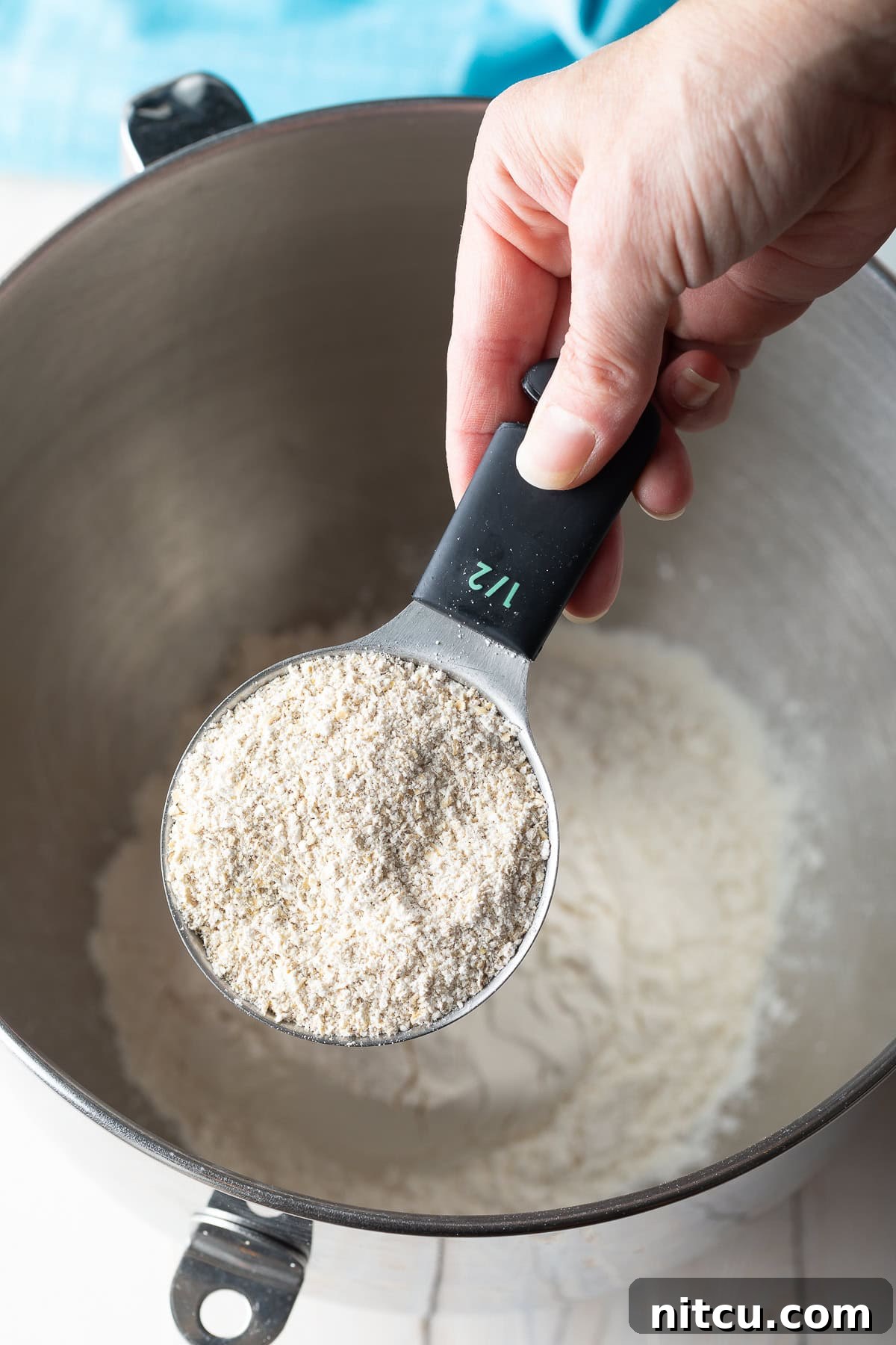 Salted Chocolate Chip Cookie Bliss 6 A hand using a measuring scoop to add oat flour to a metal mixing bowl, part of the dry ingredient combination.
