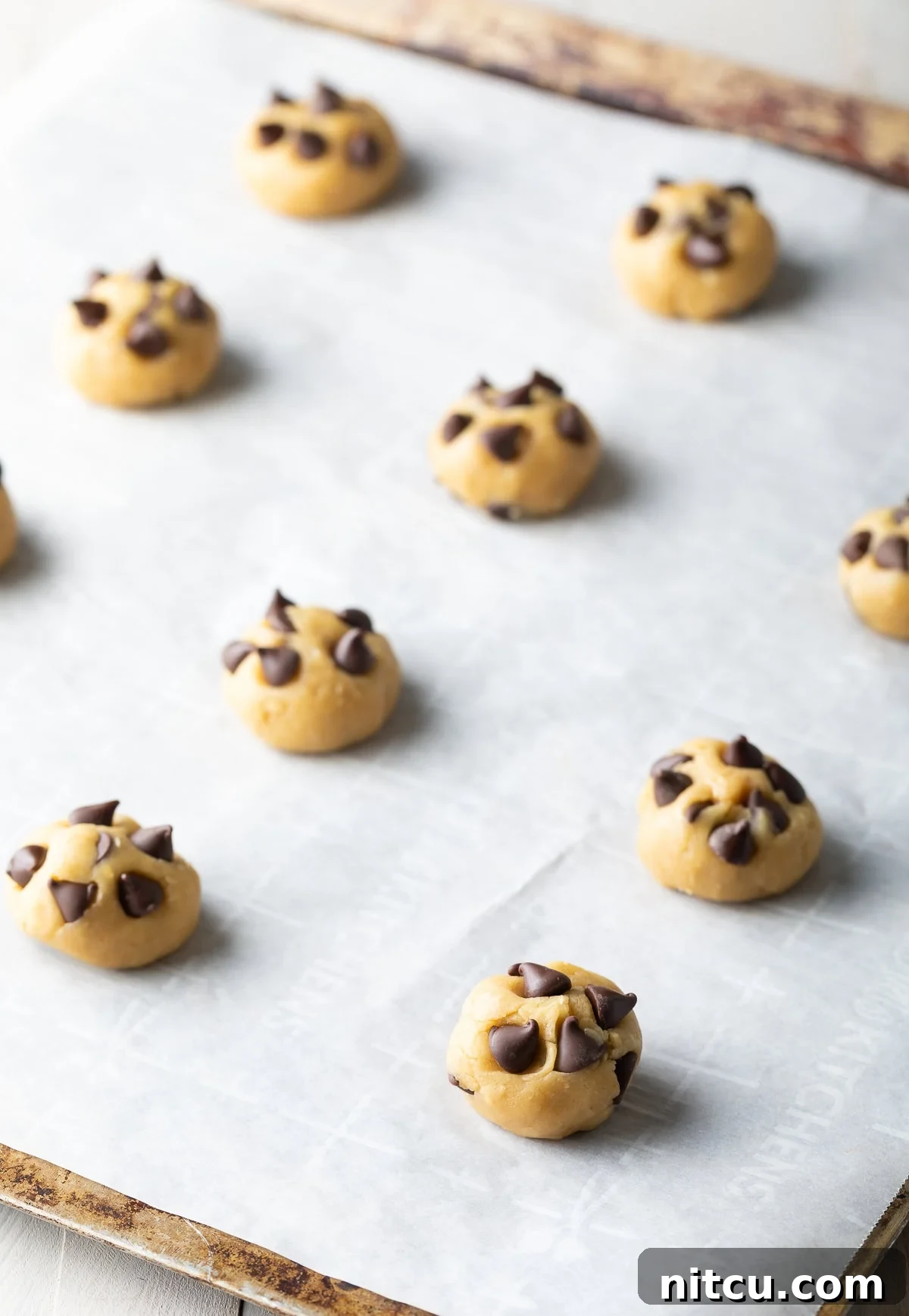 Salted Chocolate Chip Cookie Bliss 9 A dozen raw chocolate chip cookie dough balls, topped with extra chips, neatly arranged on a parchment-lined baking sheet, ready for the oven.