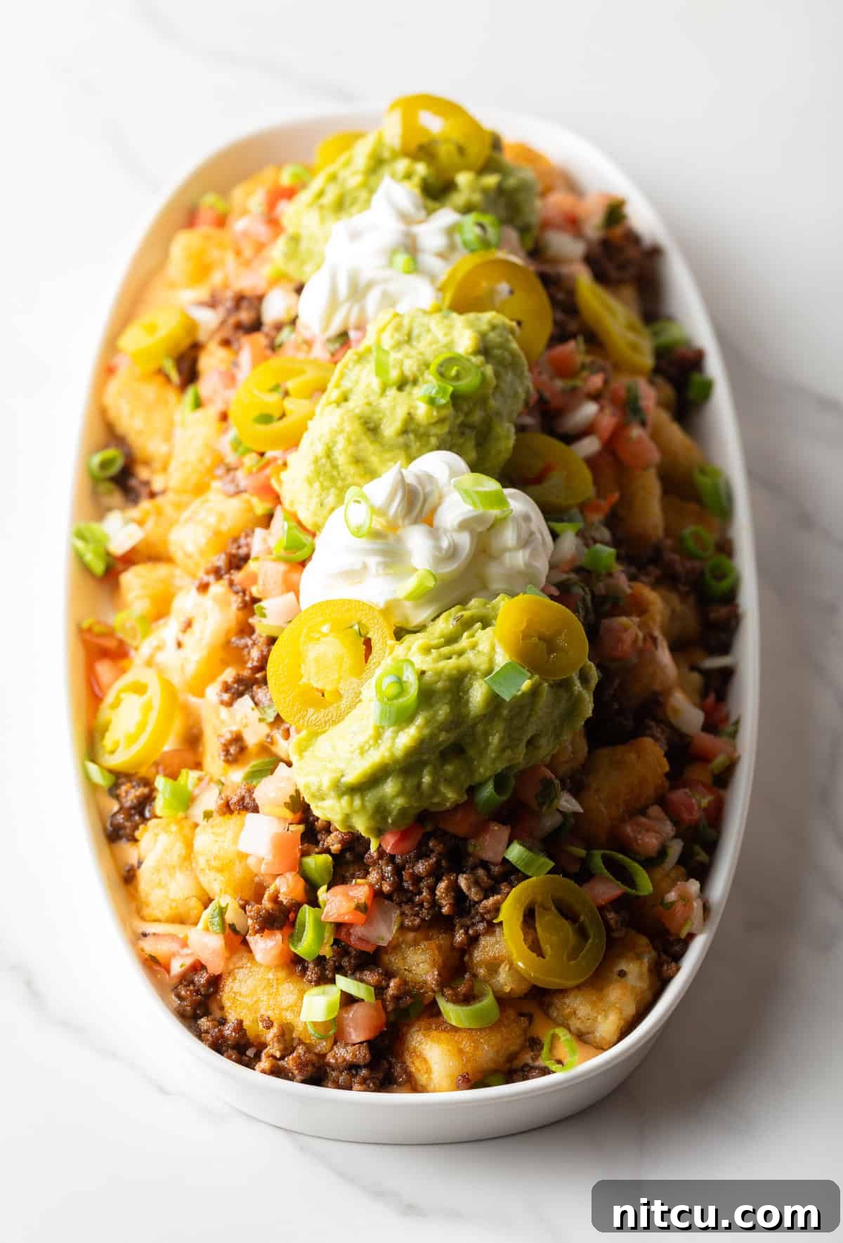 Ultimate Loaded Tots 11 Top down view of large white oval serving dish loaded with tater tot nachos with ground beef, jalapenos, salsa, cheddar cheese, guacamole and sour cream.