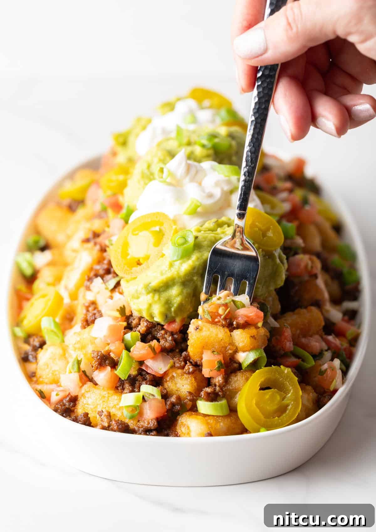 Ultimate Loaded Tots 12 Hand with a fork spearing a bite of totchos from a large white oval serving dish.