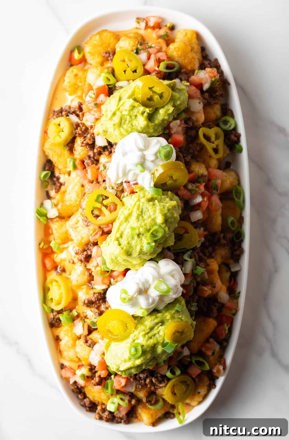 Ultimate Loaded Tots 3 Top down view of large white oval serving dish loaded with tater tot nachos with ground beef, jalapenos, salsa, cheddar cheese, guacamole and sour cream.