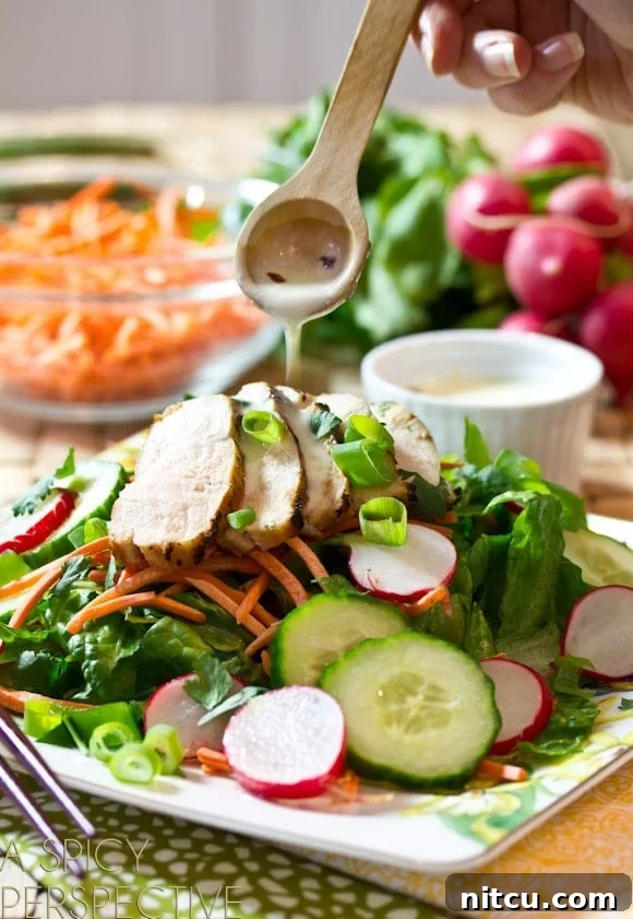 Deconstructed Banh Mi Salad 6 A close-up shot of a hand elegantly drizzling the creamy Vietnamese dressing over a vibrant plate of Banh Mi salad. The salad is loaded with crisp greens, bright pickled cucumbers and radishes, shredded carrots, and tender slices of grilled chicken breast, all awaiting the final touch of dressing.