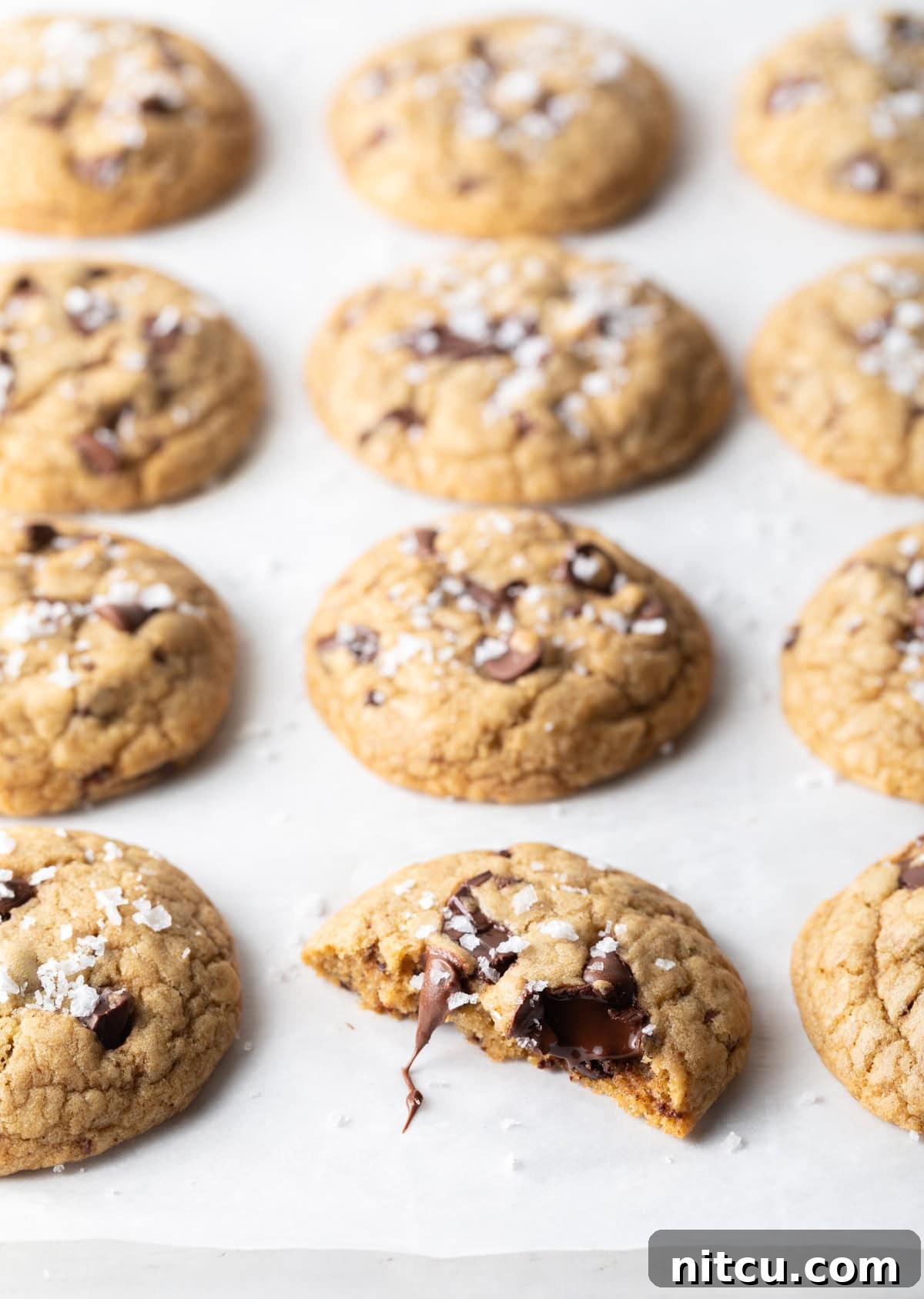 A top-down shot of a baking sheet filled with golden-brown chocolate chip cookies. One cookie in the foreground is broken in half, revealing its soft, chewy interior and melty chocolate.