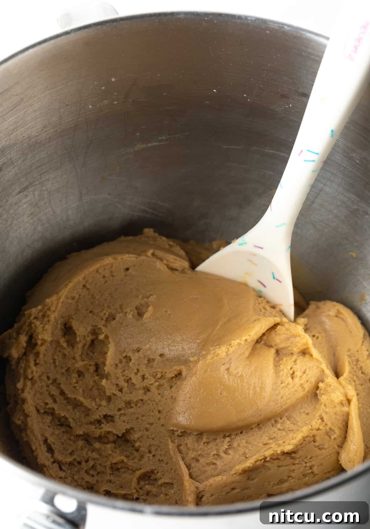 Rich brown sugar cookie dough nestled in a gleaming metal mixing bowl, with a rubber spatula resting beside it.