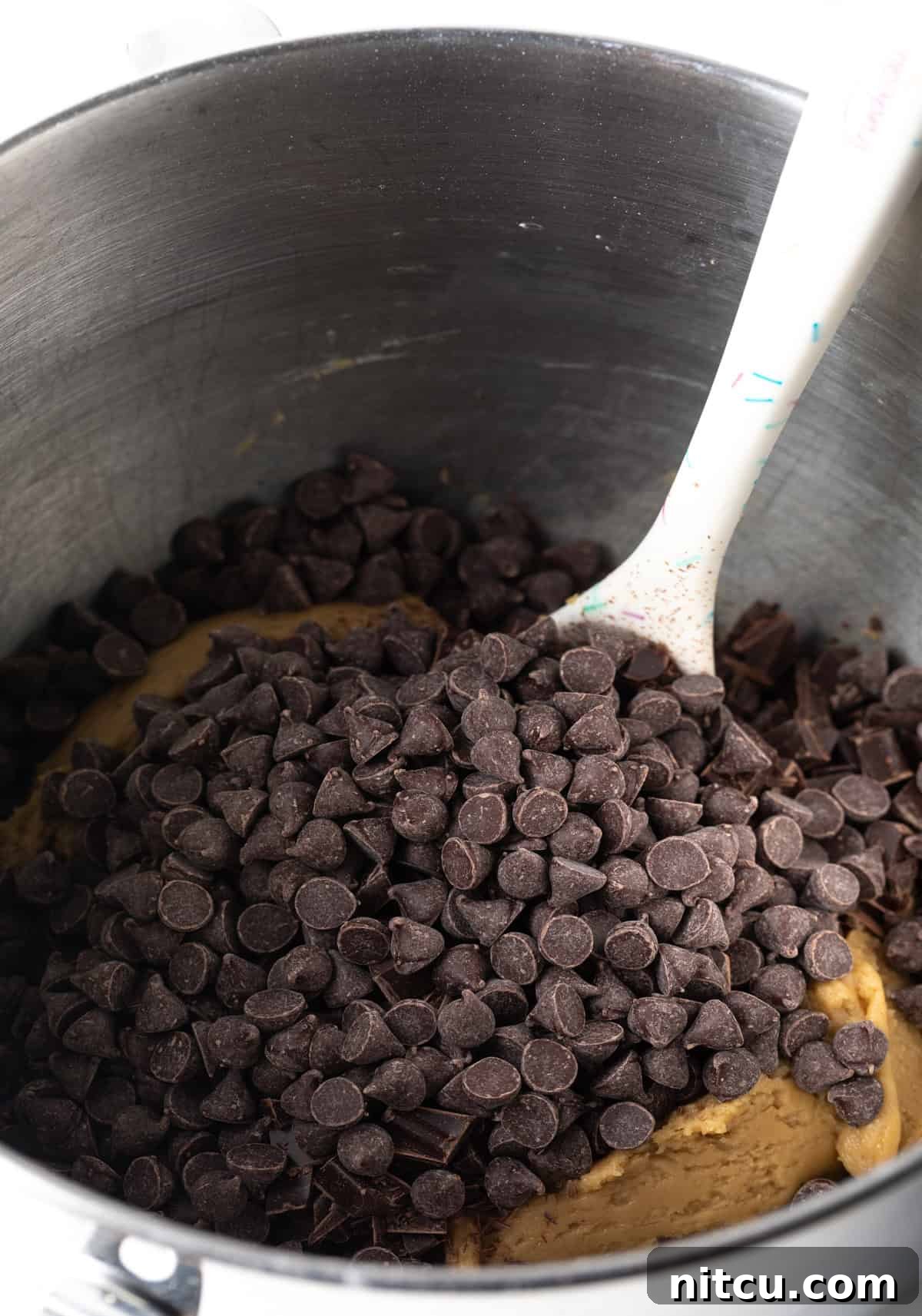 Semi-sweet chocolate chips being added to the rich brown sugar cookie dough in a metal mixing bowl.
