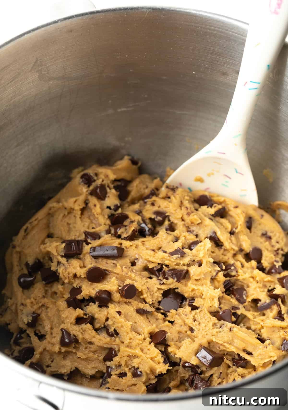 A spatula gently stirring chocolate chunks into the thick, luscious brown sugar cookie dough, ensuring even distribution.