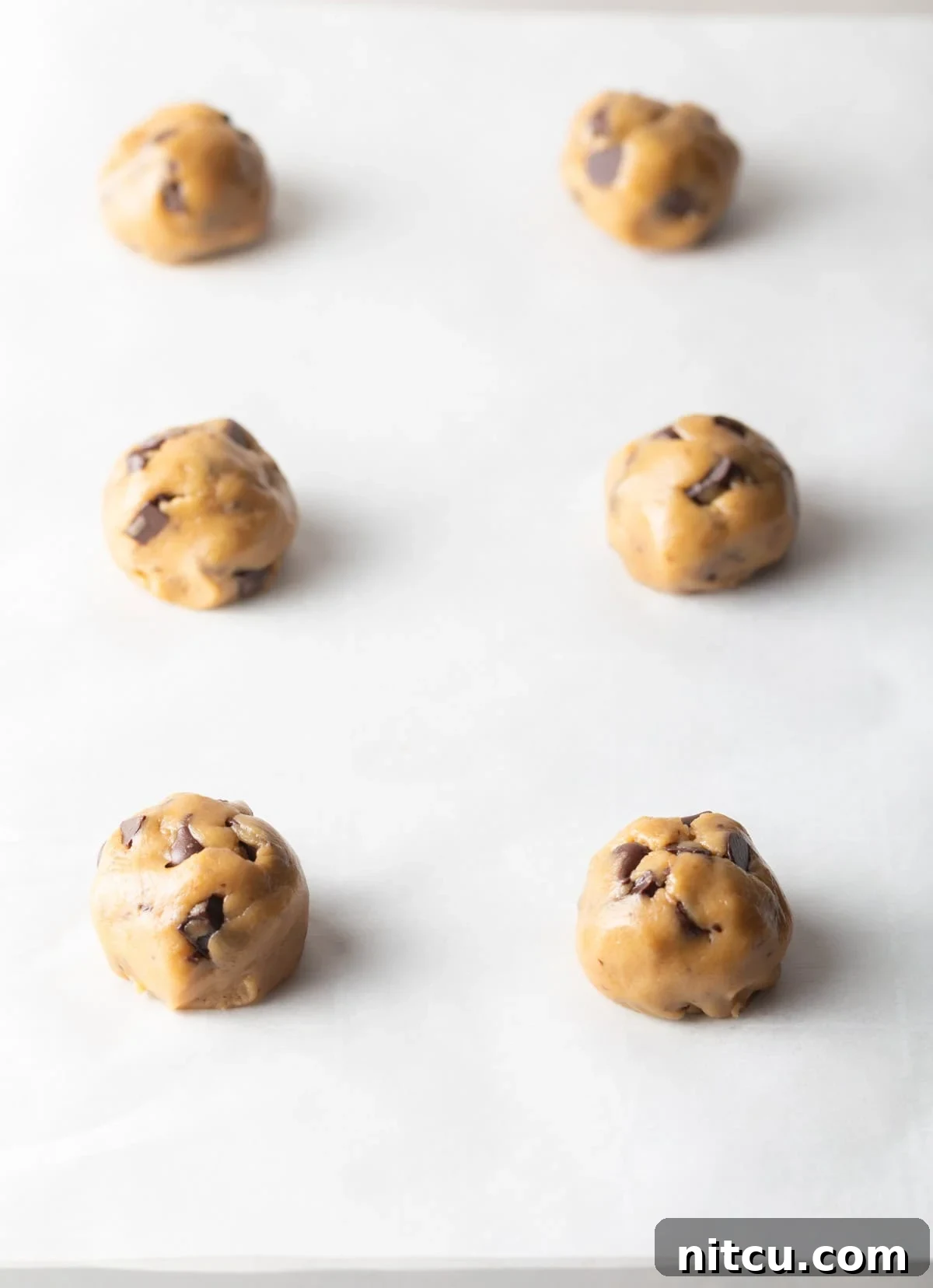 Six perfectly scooped and rolled cookie dough balls, strategically placed on a baking sheet, ready for the oven.