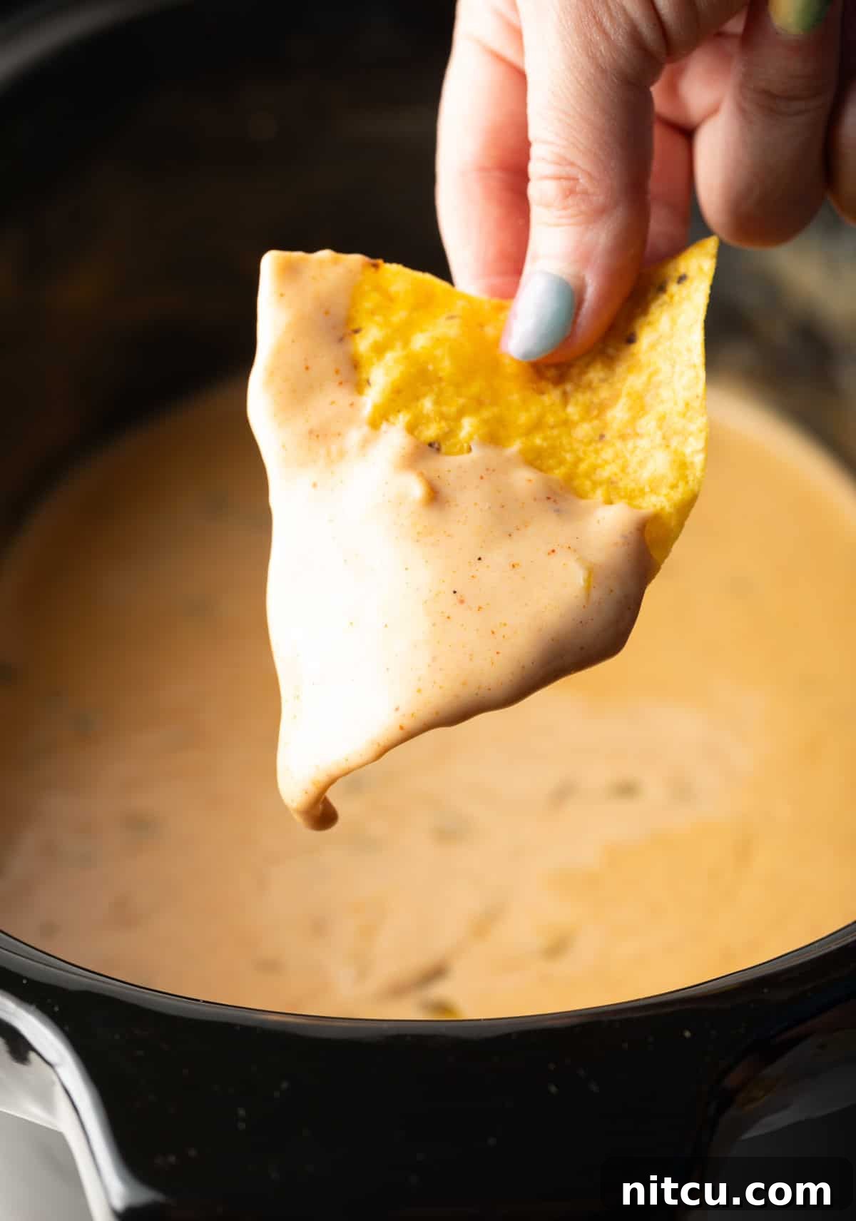 Ultimate Crock Pot Queso Dip 5 A hand holding a tortilla chip that's been dipped in the crock pot queso recipe hovered over the crock pot.