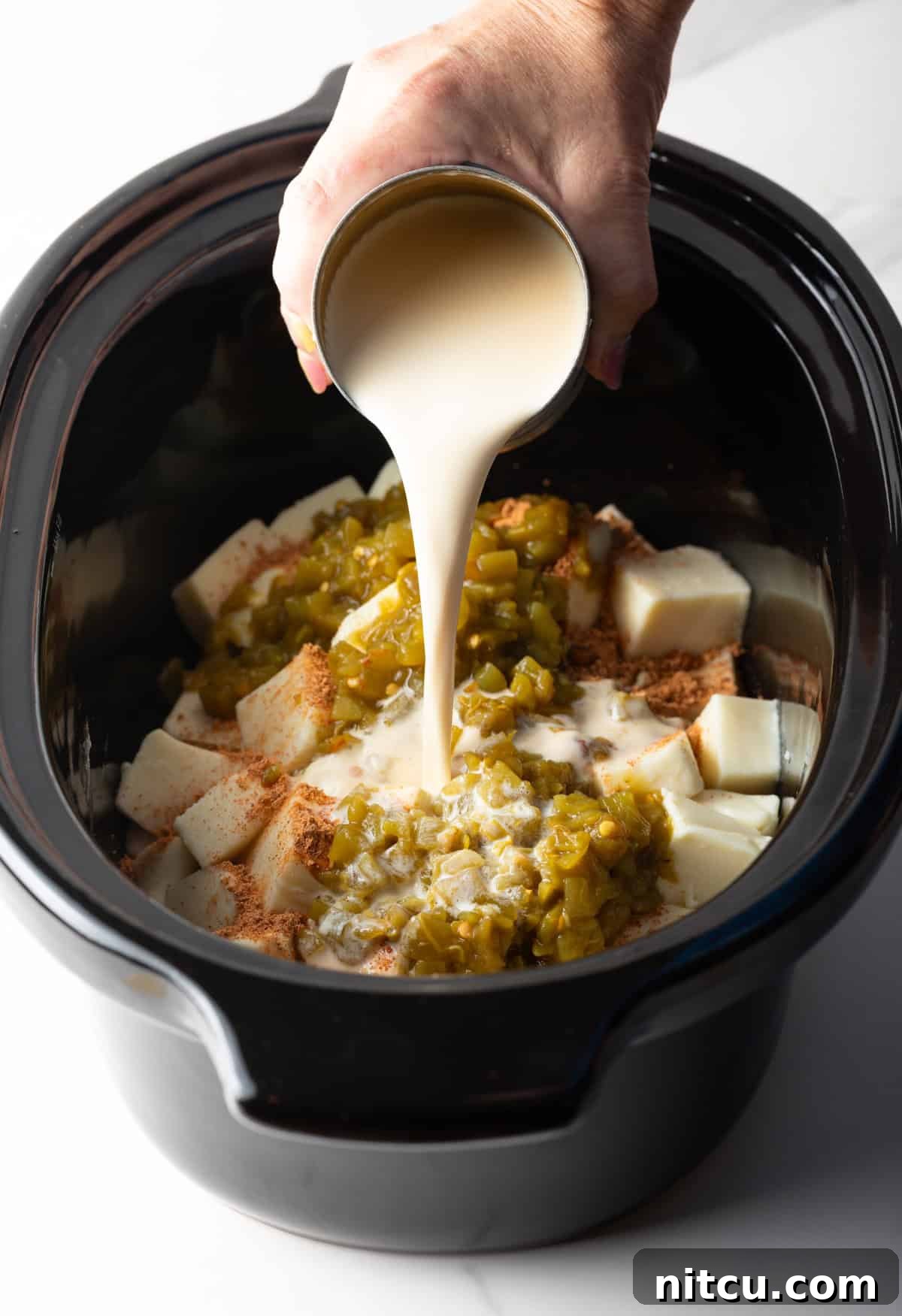 Ultimate Crock Pot Queso Dip 7 Crock pot queso ingredients assembled in a black crock pot with evaporated milk being poured over it.