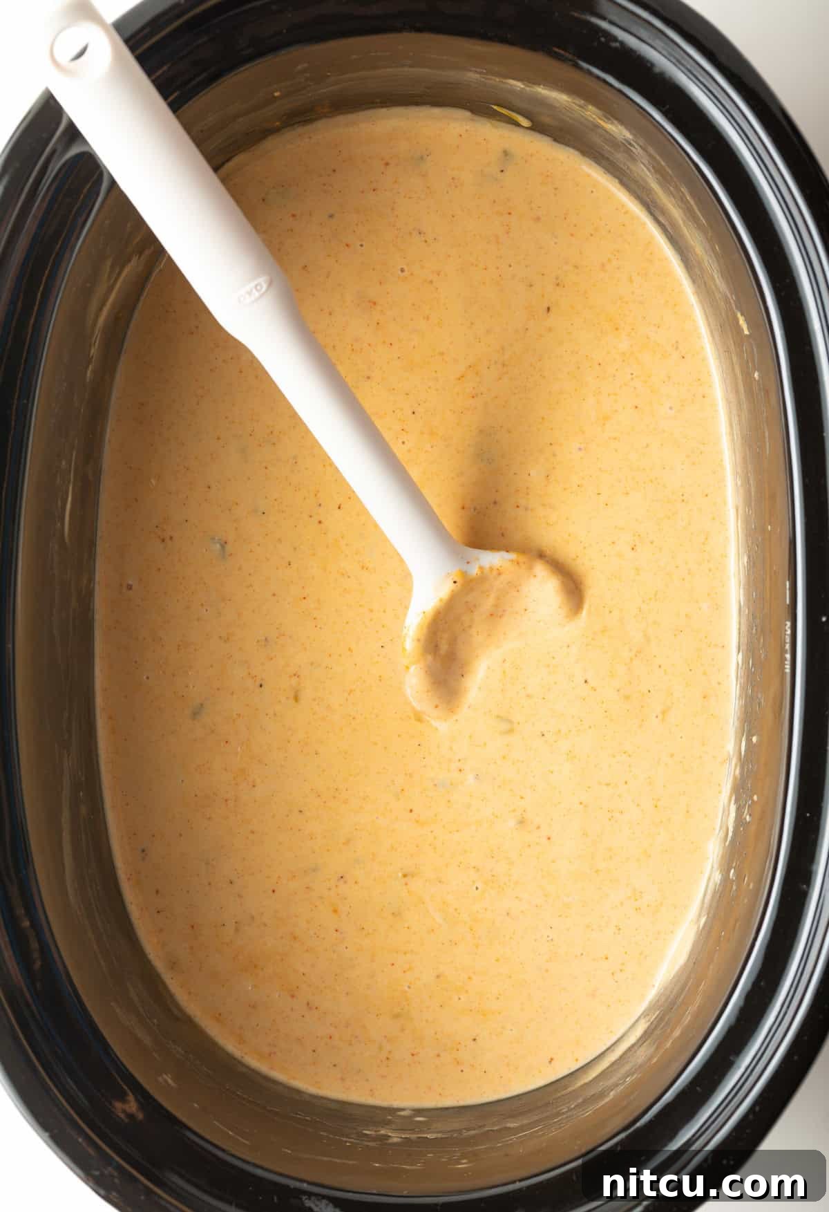 Ultimate Crock Pot Queso Dip 8 Black slow cooker filled with queso dip with a white spoon stirring it.