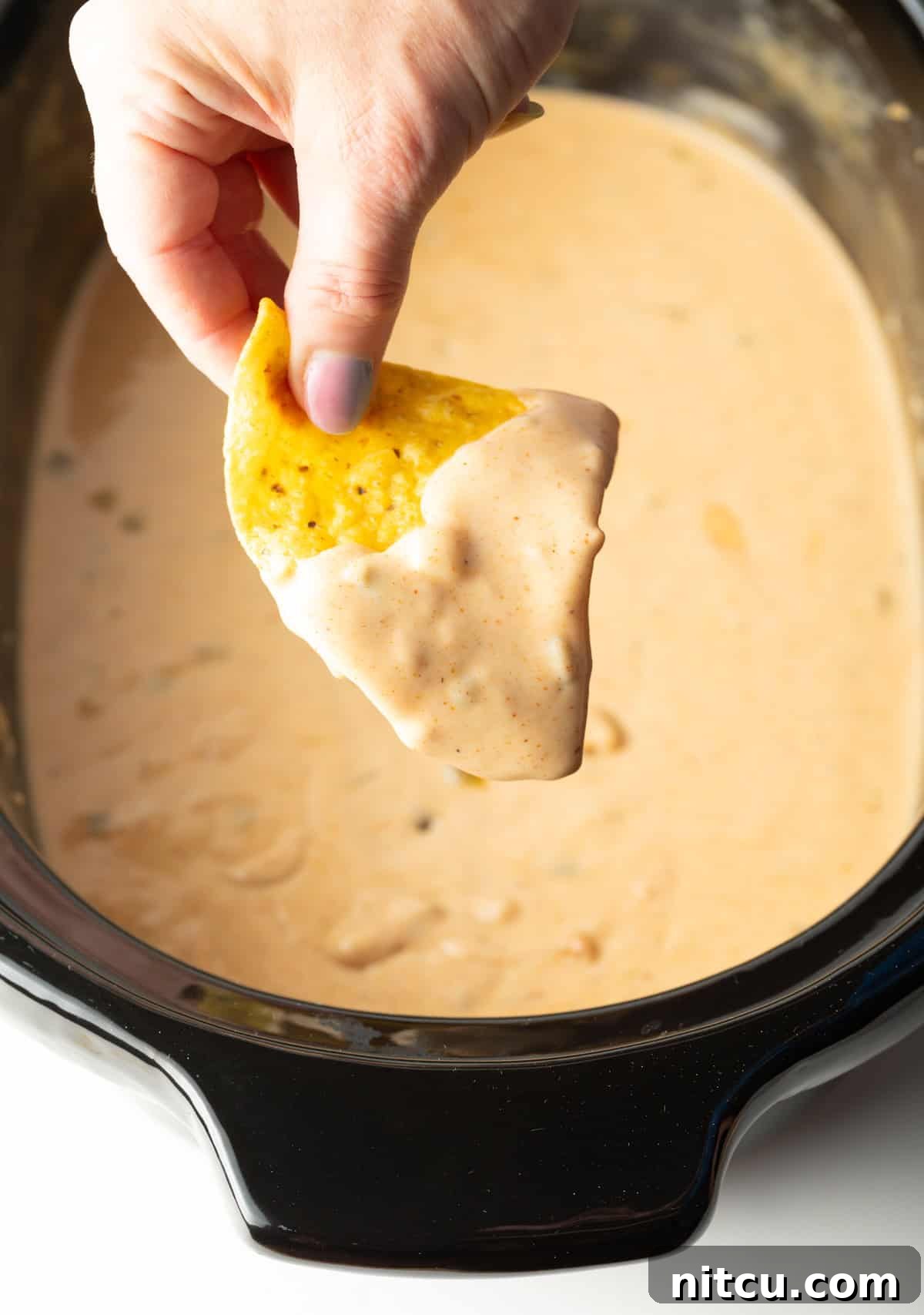 Ultimate Crock Pot Queso Dip 9 A hand holding a tortilla chip that's been dipped in crock pot queso hovered over the crock pot.