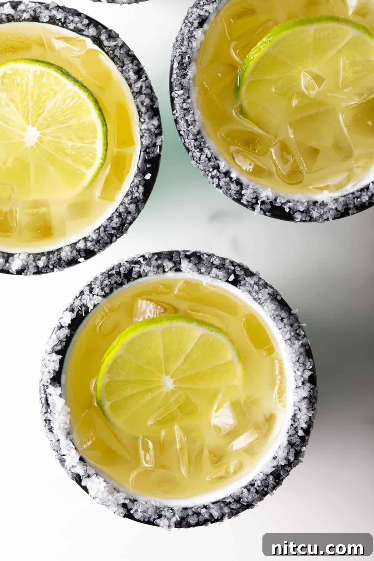 Zero-Proof Margarita 3 Top down view of 3 white and green-striped cups with margaritas on the rock, with salt around the rim and a slice of lime in the drink.