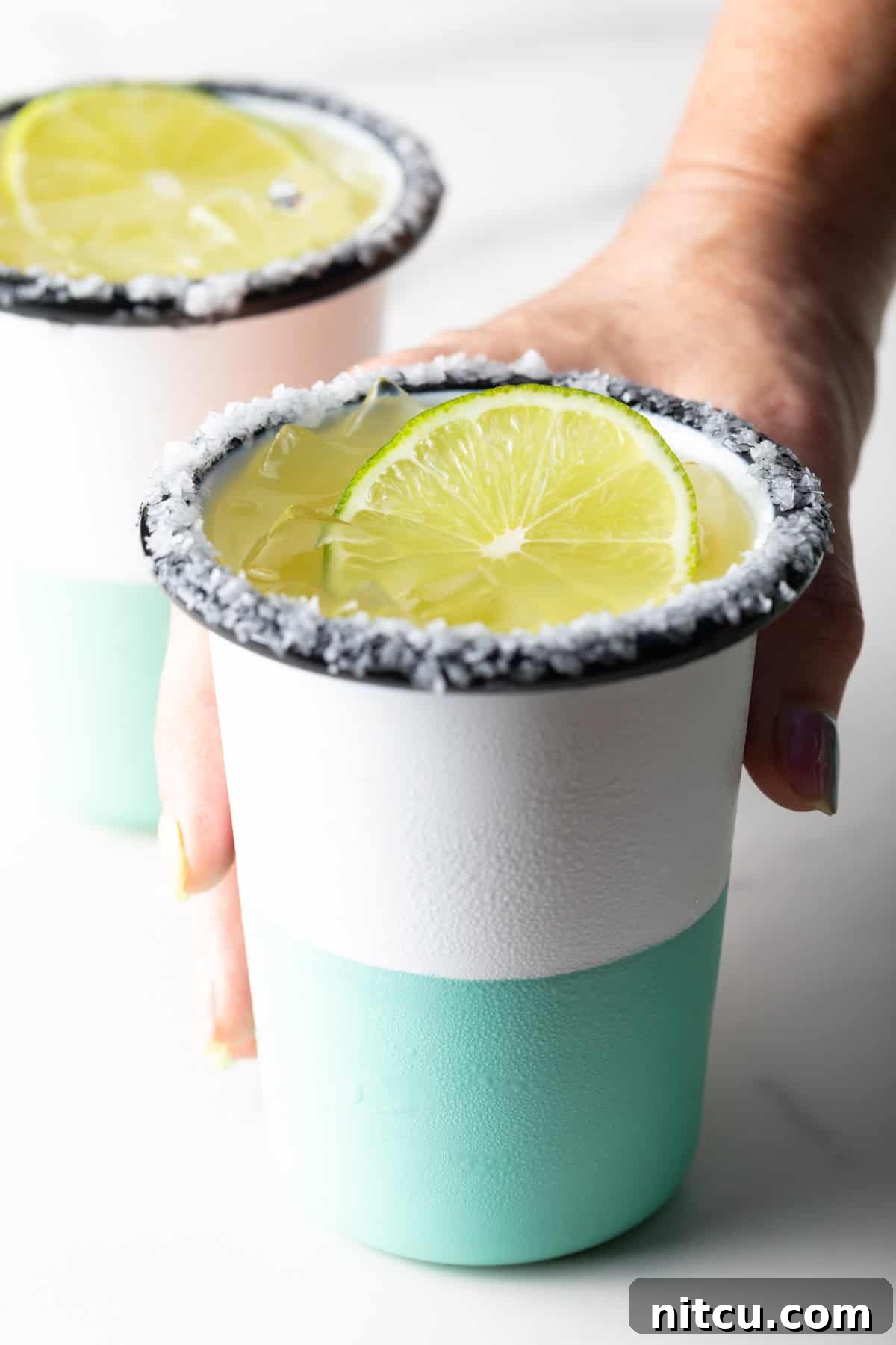 Zero-Proof Margarita 9 Hand grabbing a white and green-striped cup with salt around the rim and slice of lime in the drink.
