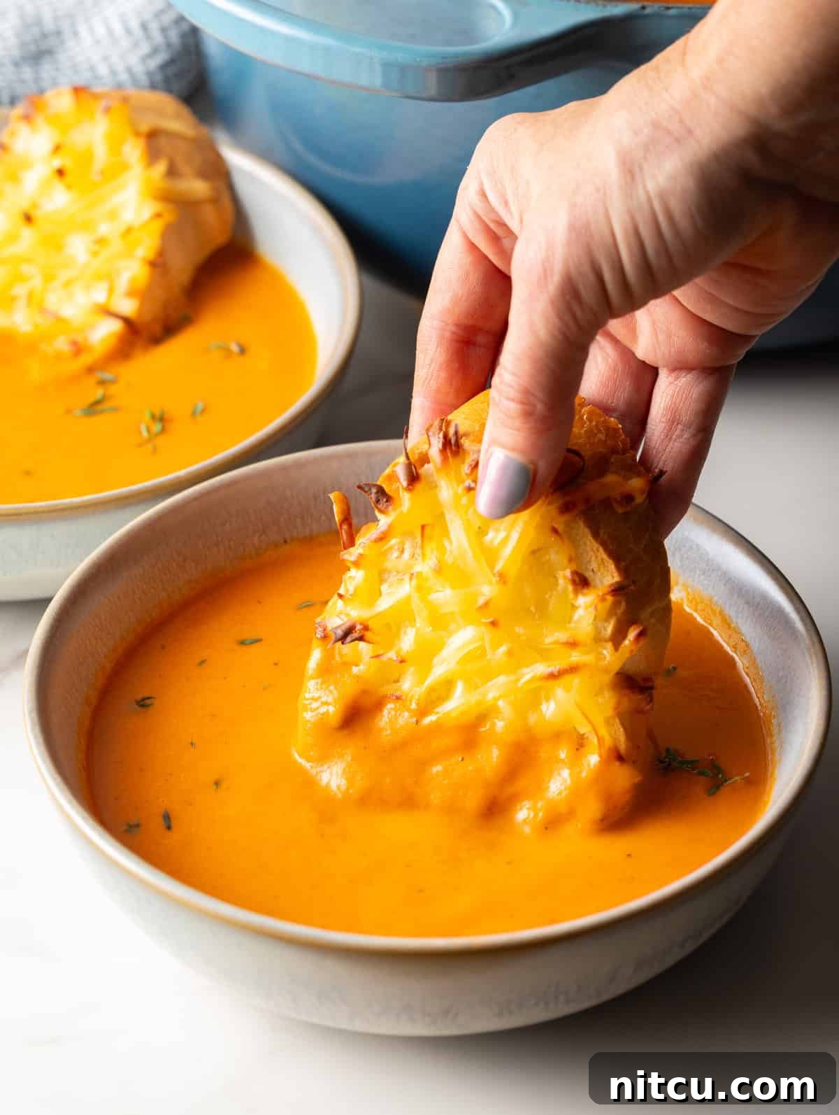 Smoky Red Pepper Bisque 2 A hand holding cheesy Gouda baguette toast, freshly baked, dipping it into a bowl of vibrant, creamy roasted red pepper soup.