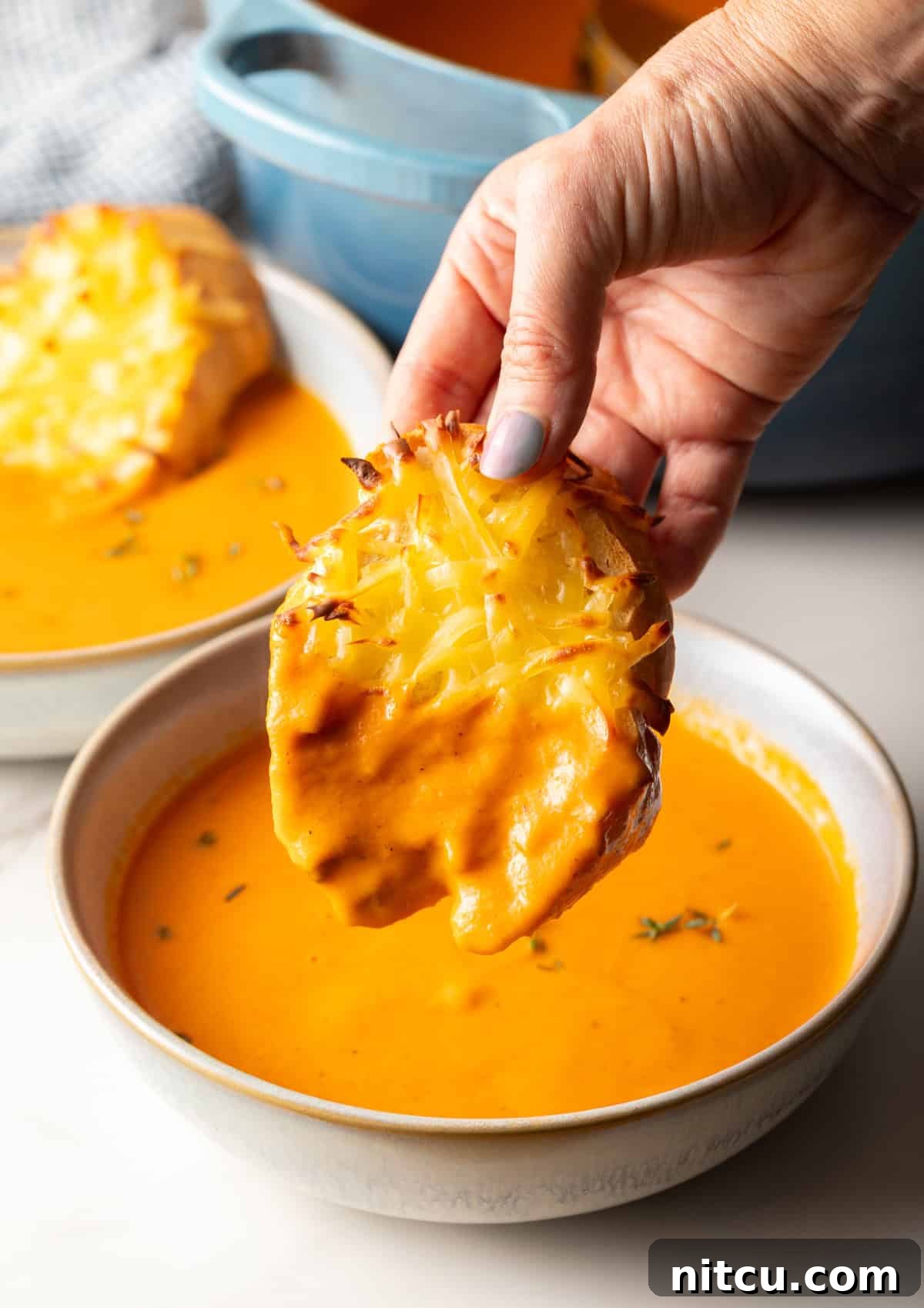 Smoky Red Pepper Bisque 5 A close-up shot of a hand dipping a piece of bubbly, golden Gouda cheese toast into a rich, creamy bowl of roasted red pepper Gouda soup.