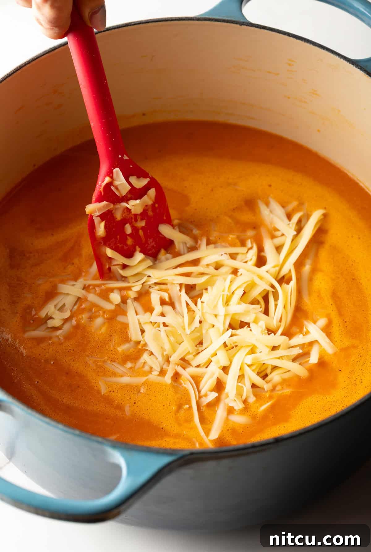 Smoky Red Pepper Bisque 10 A generous handful of freshly shredded smoked Gouda cheese being stirred into a steaming pot of rich, creamy red pepper soup, ready to melt into a velvety texture.