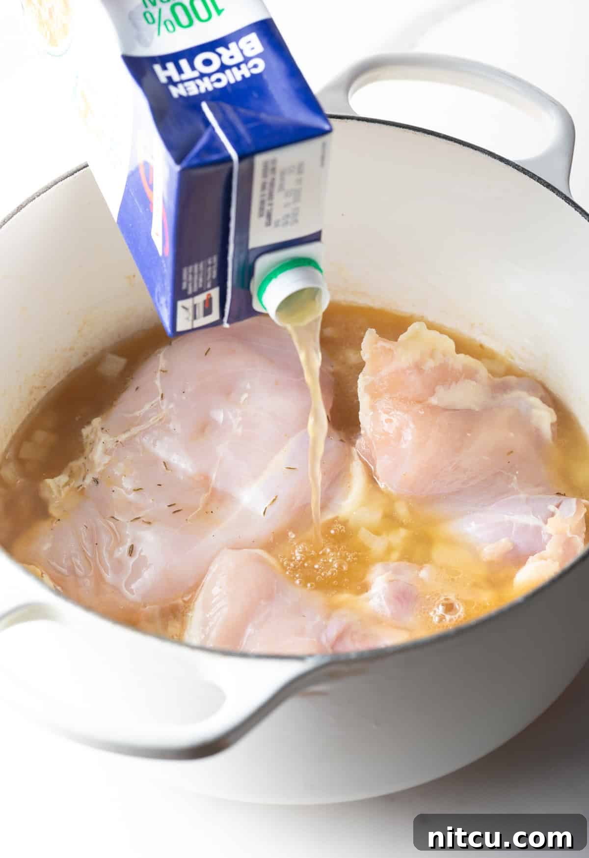 Adding chicken broth from a carton to a soup pot with raw chicken breasts.
