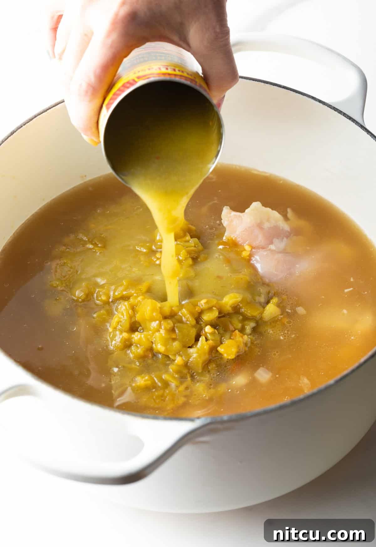 Pouring verde enchilada sauce from a can into a white pot of soup.