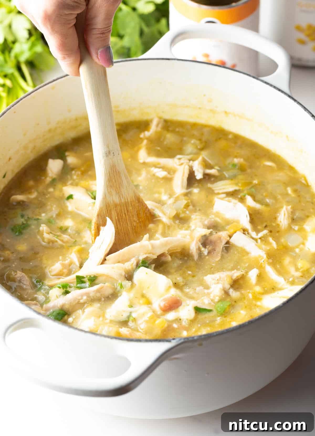 Hand using a large wooden spoon to stir shredded chicken into a large white cooking pot.