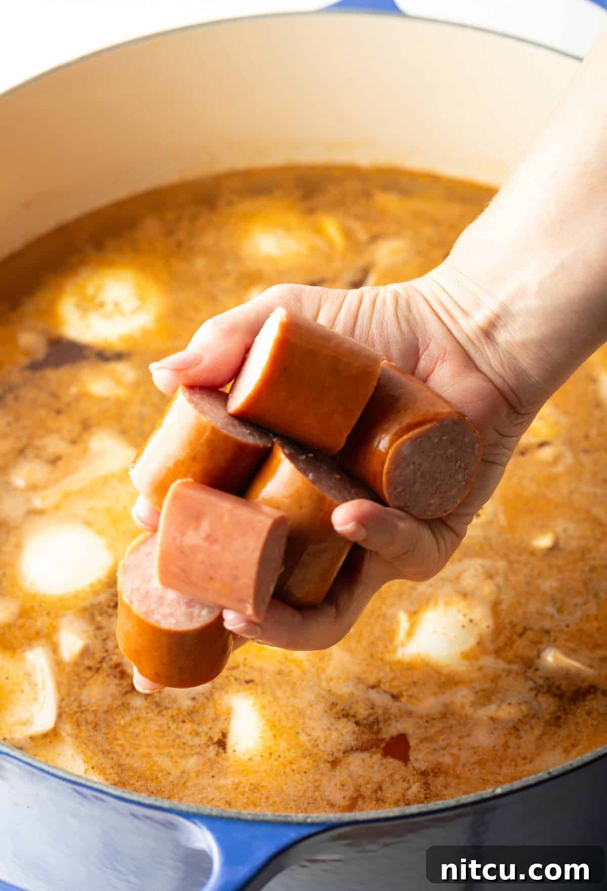 Coastal Bounty Boil 6 A hand carefully placing thick slices of kielbasa sausage into a steaming pot, joining the onions, potatoes, and fragrant Old Bay broth, simmering for the authentic Low Country Boil recipe.