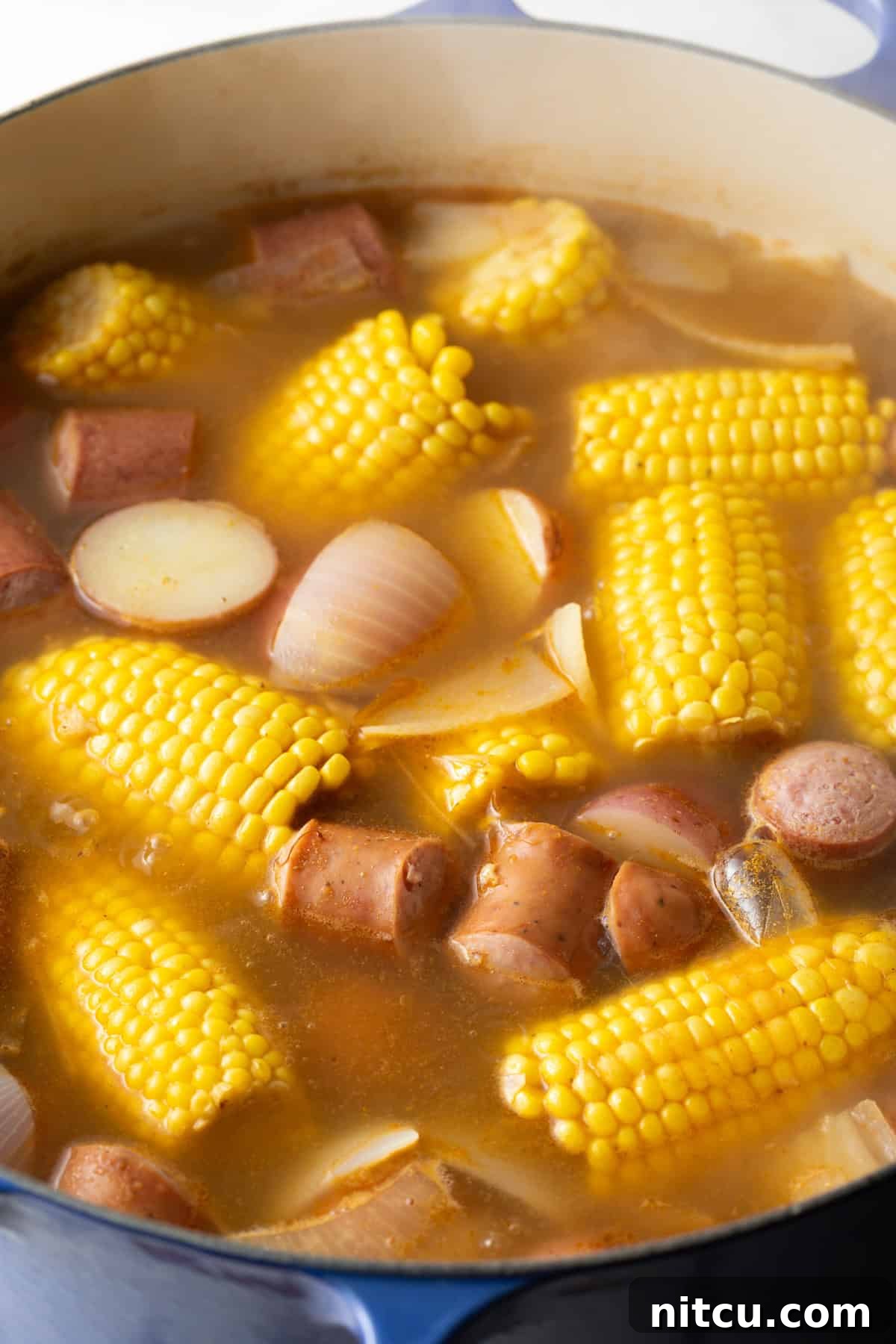 Coastal Bounty Boil 7 A bubbling pot brimming with the rich, Old Bay spiced broth, featuring vibrant yellow corn on the cob, tender red potatoes, savory onions, and smoky sausage, all simmering together.