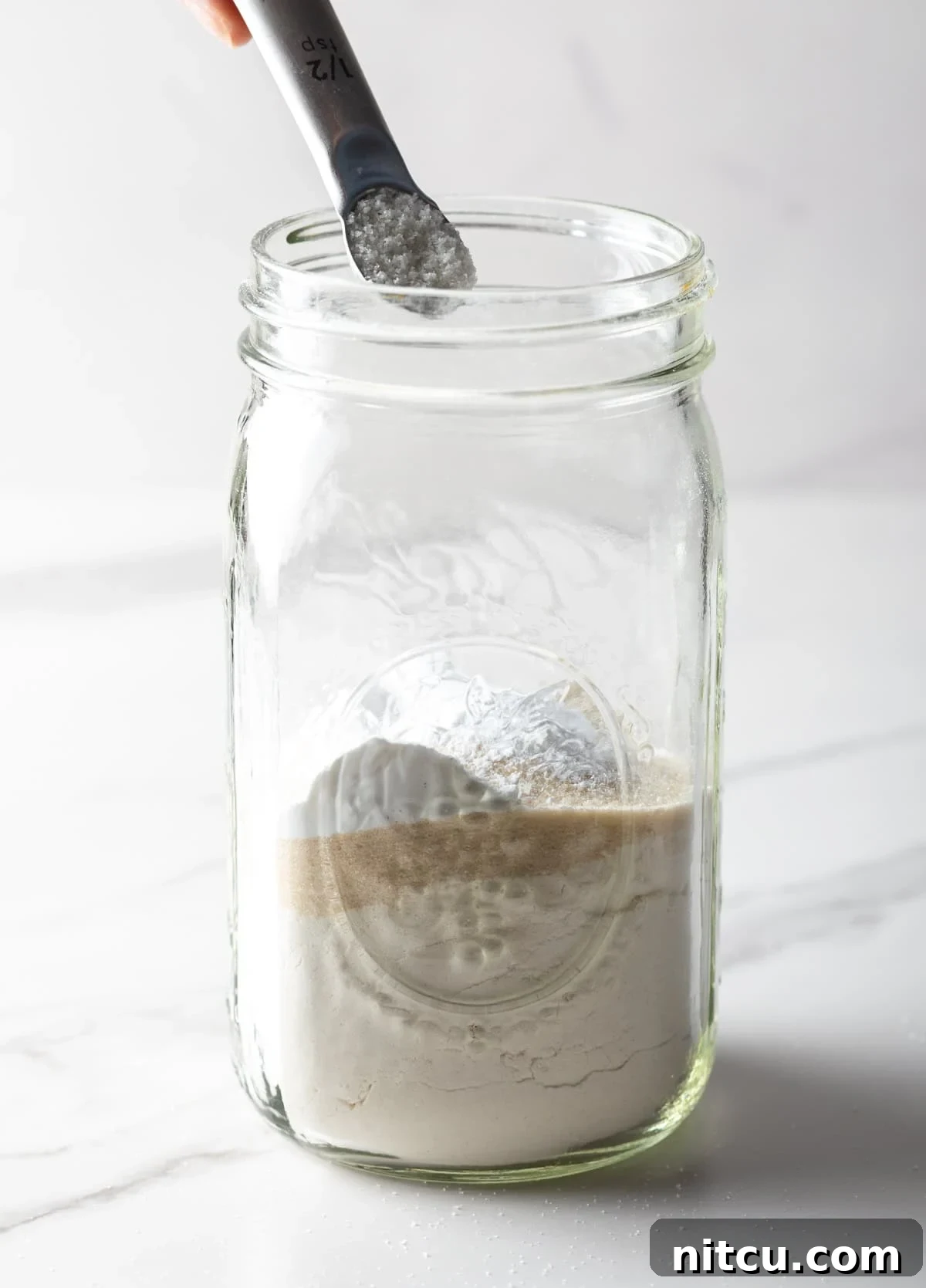A measuring spoon scooping baking soda into a glass jar of homemade pancake mix.