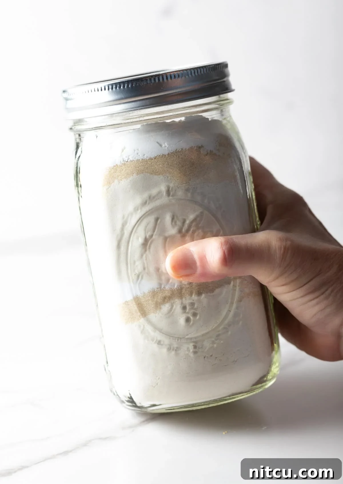 A hand holding a glass jar filled with homemade pancake mix ingredients, ready to be shaken.