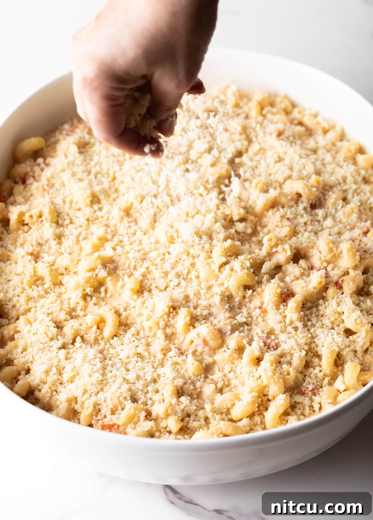Big Easy Mac and Cheese 10 Breadcrumbs and grated parmesan are being sprinkled over the Cajun mac and cheese in a white baking dish.