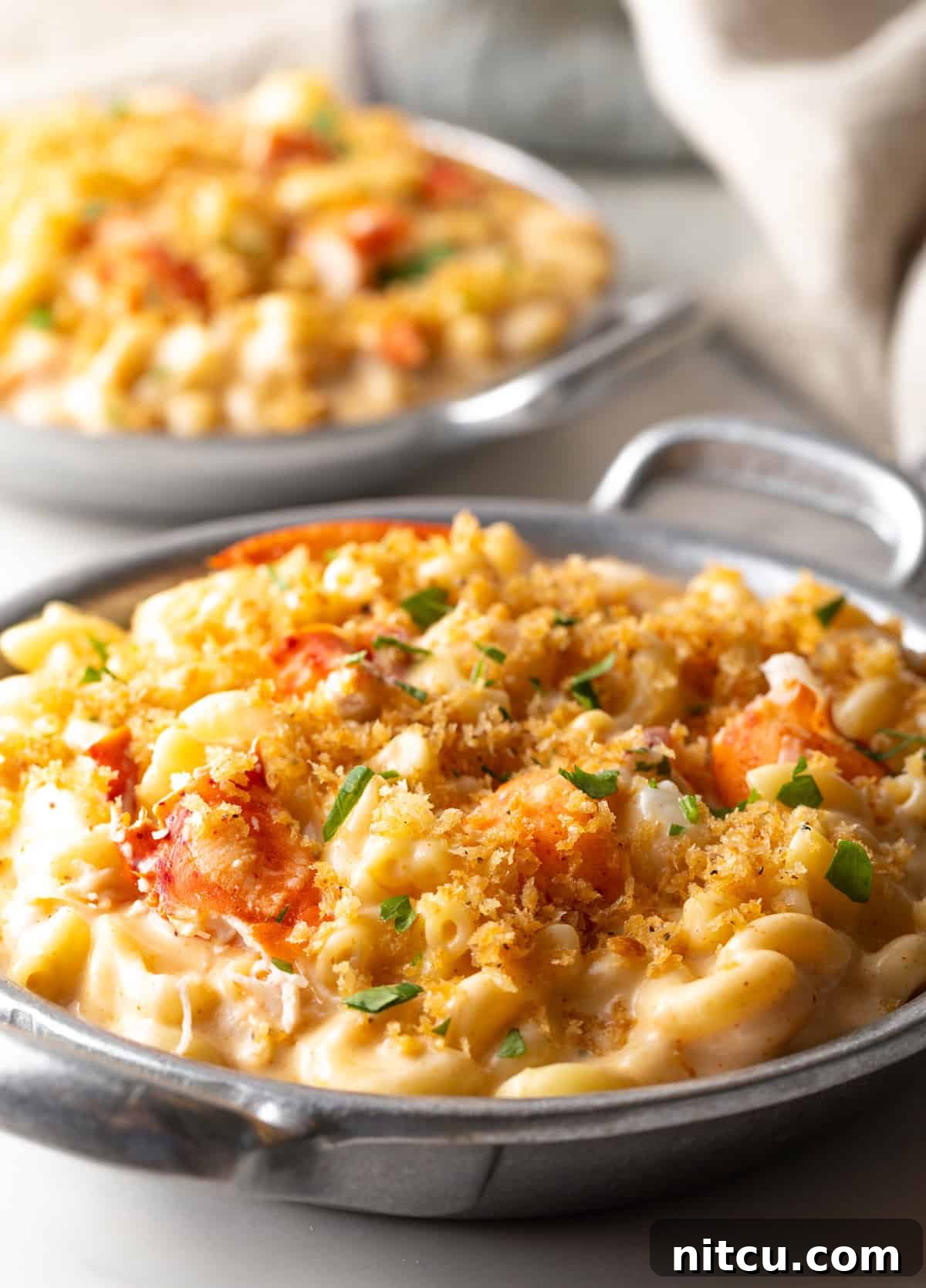 Decadent Lobster Mac and Cheese 3 Side view of a metal bowl filled with a lobster mac and cheese recipe topped with breadcrumbs and parsley.