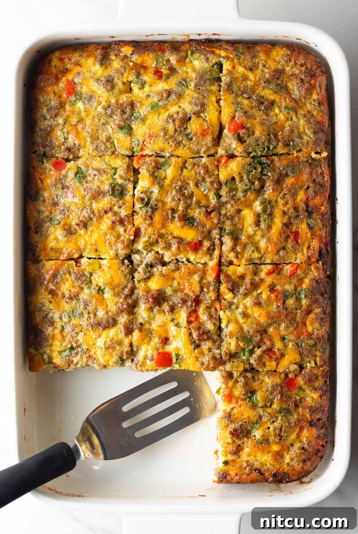 Savory Morning Sausage Bake 2 Pan of egg casserole with sausage and vegetables. A spatula is in the pan, with square portions of the casserole removed.