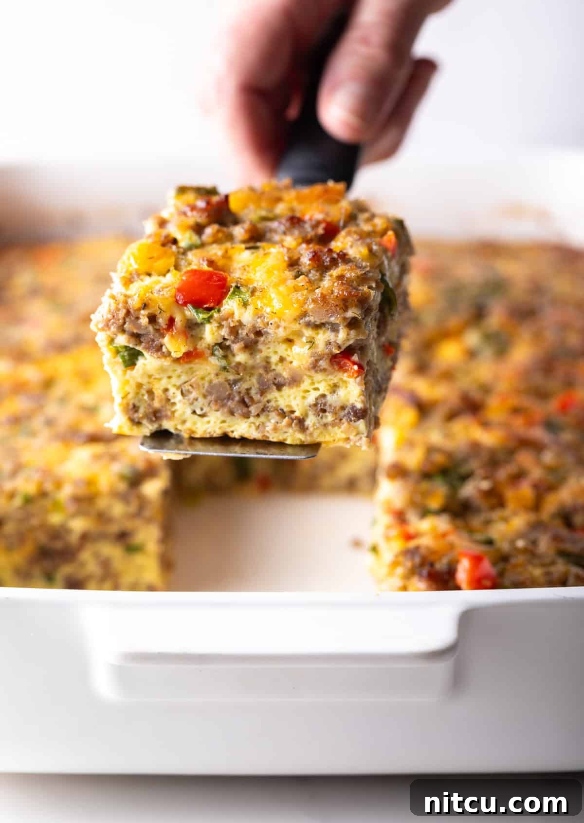 Savory Morning Sausage Bake 3 Pan of egg casserole with sausage and vegetables. A hand is holding a spatula above the pan, with a square portion.