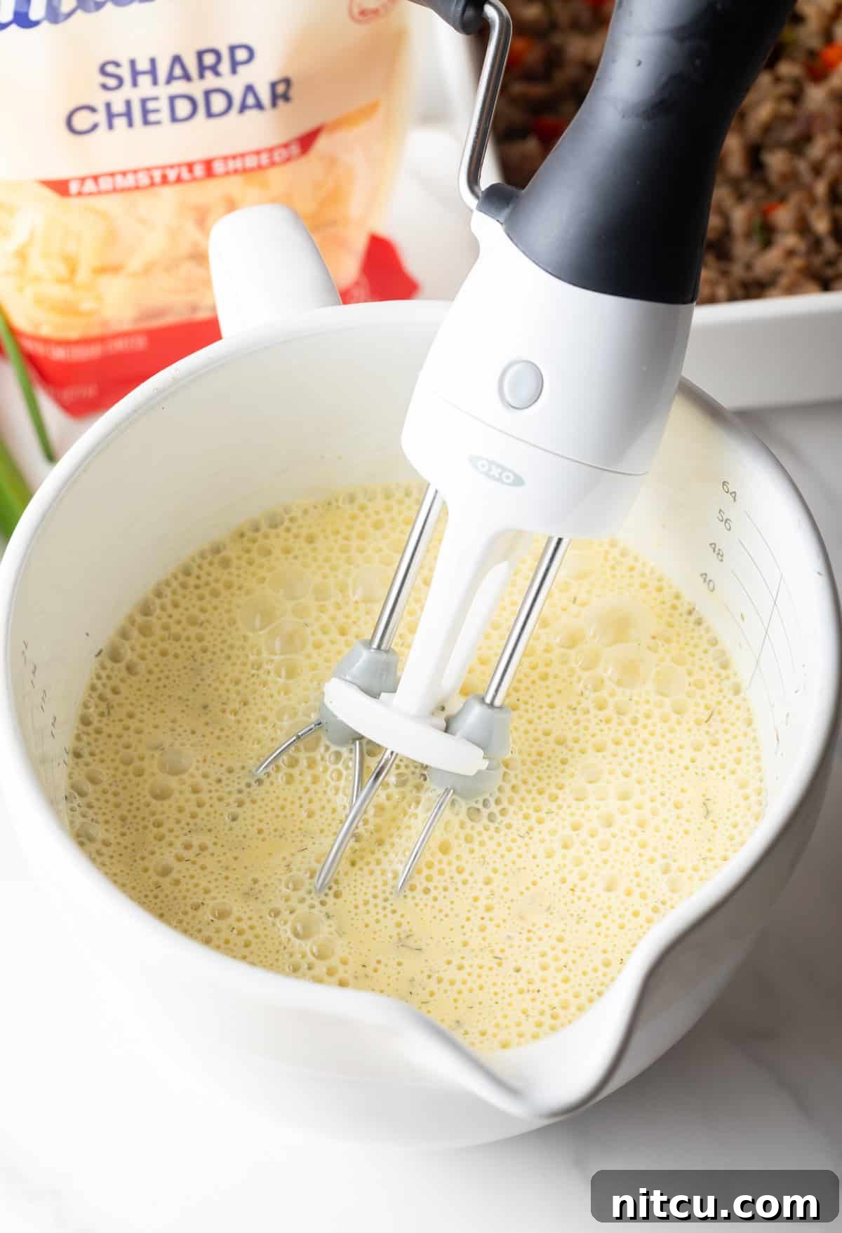 Savory Morning Sausage Bake 9 Hand mixer blending eggs and spices in a white measuring pitcher.