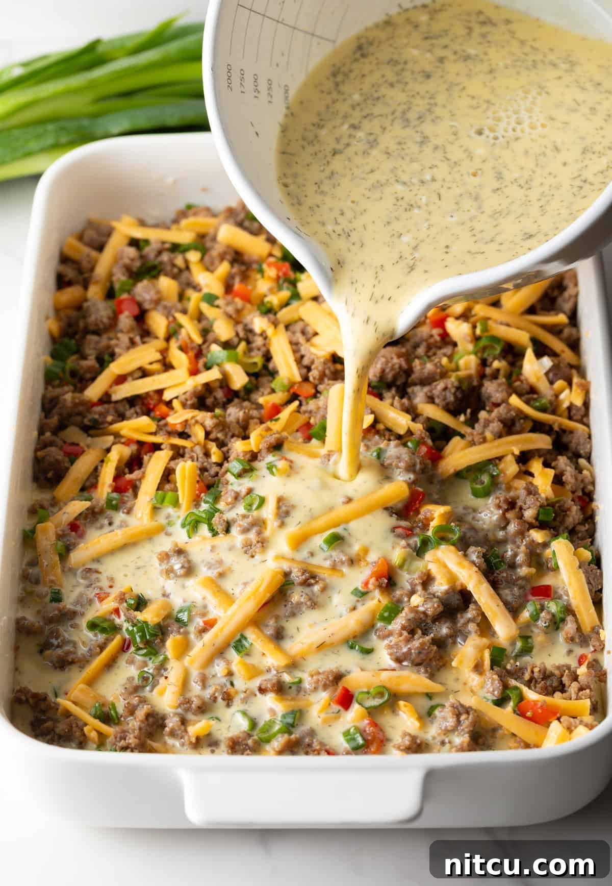 Savory Morning Sausage Bake 10 Pouring egg mixture over shredded cheese and sausage in a white rectangle baking dish.