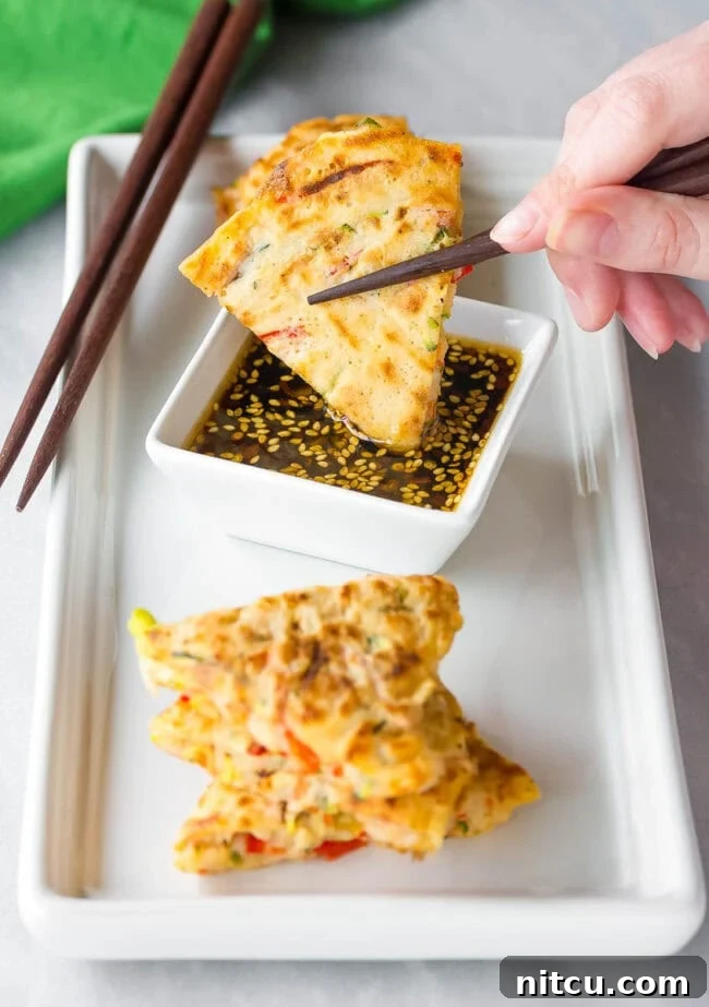 Korean Vegetable Pancake Dipping Sauce A hand is gently dipping a fresh, crispy Korean pancake wedge into a small bowl of spicy soy dipping sauce, highlighting the perfect pairing.