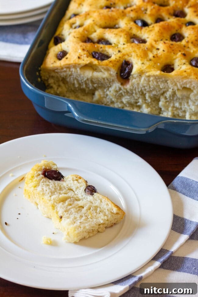 Golden Herb & Olive Focaccia 4 A loaf of focaccia is in a blue baking dish in the background. In the foreground is a white plate with a slice of fresh baked bread studded with olives.