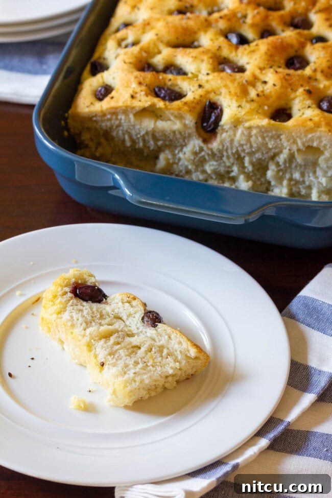 Golden Herb & Olive Focaccia 10 A loaf of focaccia is in a blue baking dish in the background. In the foreground is a white plate with a slice of fresh baked bread studded with olives.