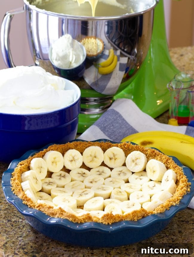 Velvet Banana Indulgence 5 Freshly sliced banana pieces being arranged onto a baked graham cracker crust in a blue pie plate.