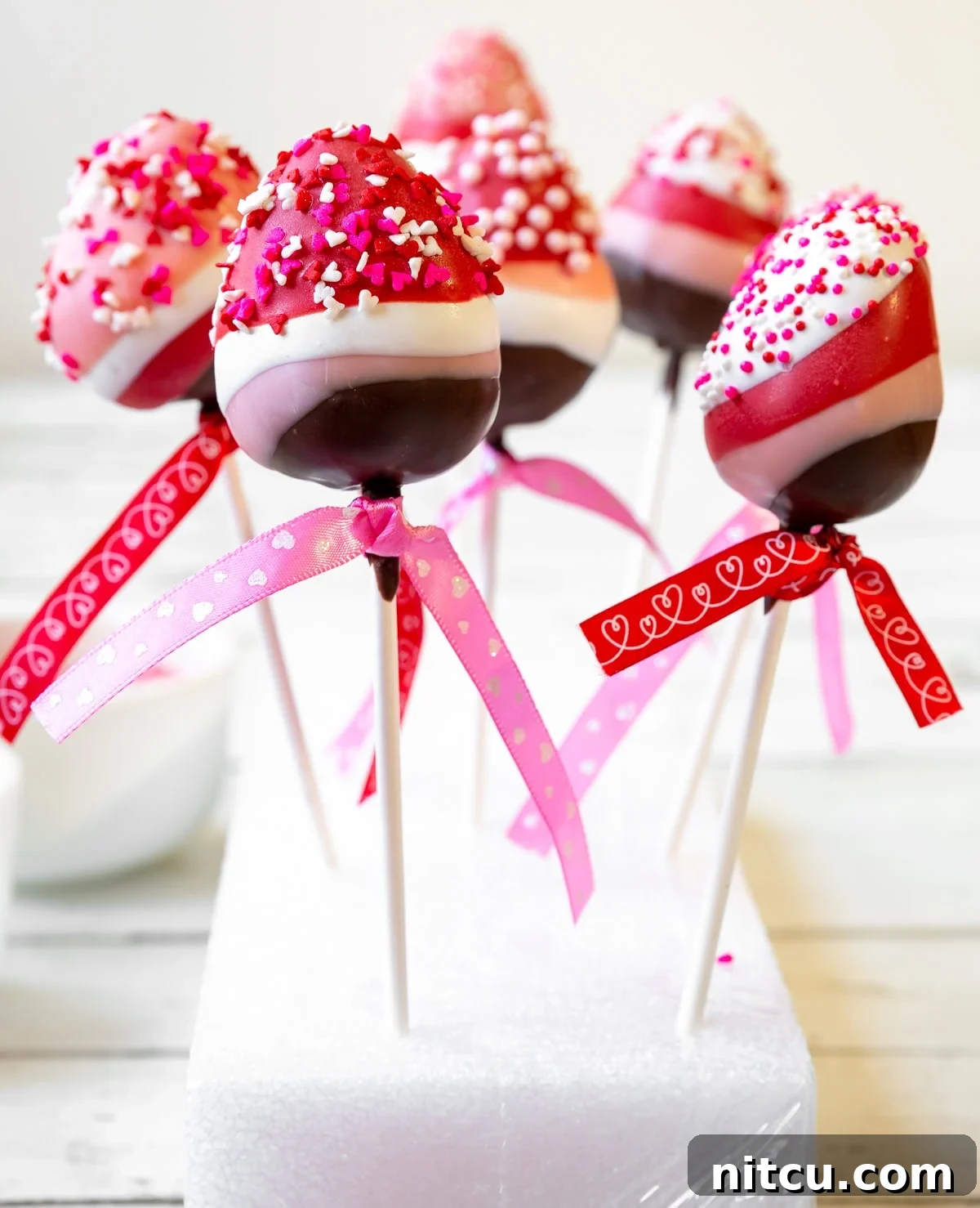 Gourmet Chocolate Strawberry Pops 2 Six vibrant chocolate-covered strawberry lollipops, featuring layers of rich dark chocolate, sweet pink, crisp white, and passionate red coatings, all adorned with festive pink and white Valentine's Day sprinkles.