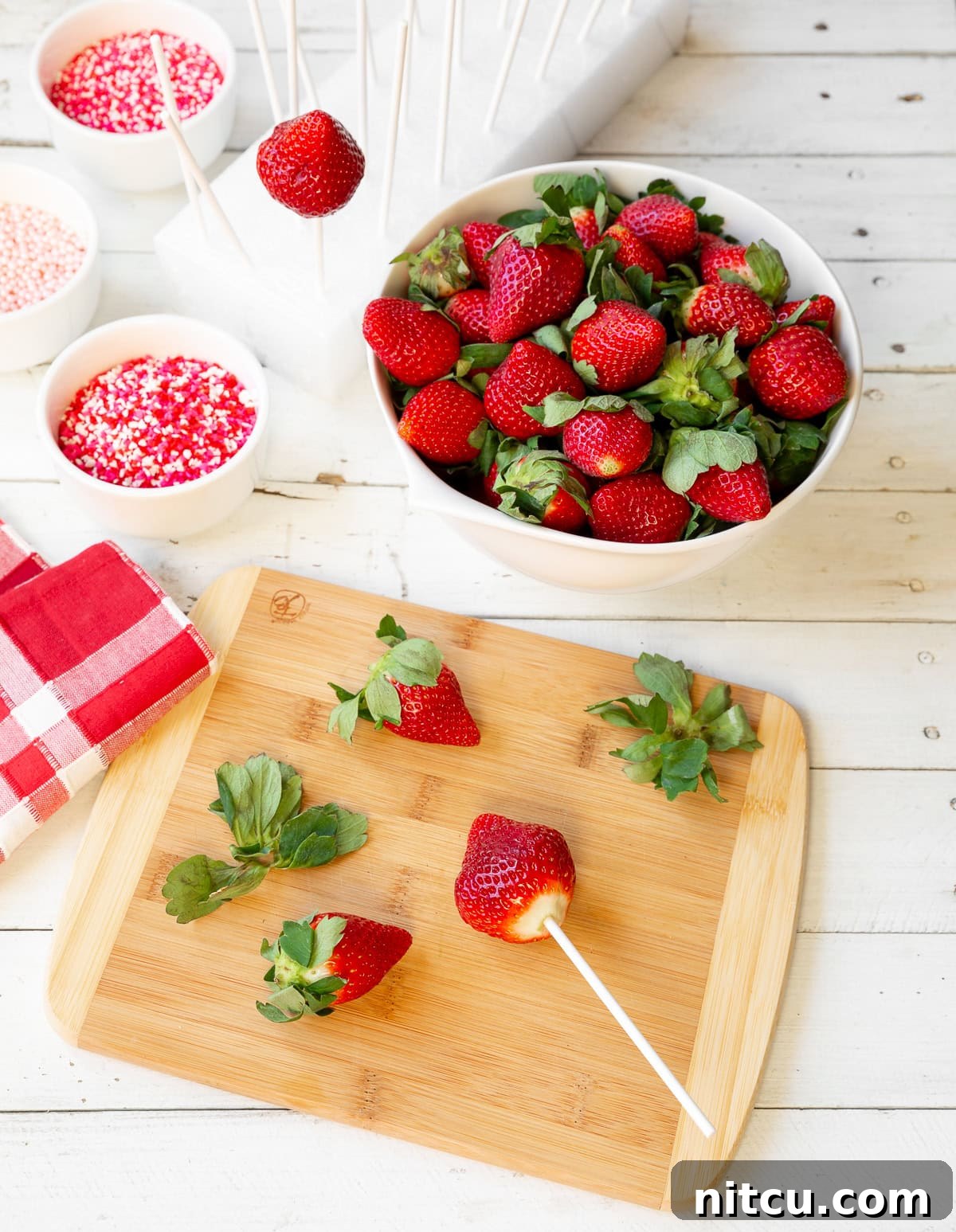 Gourmet Chocolate Strawberry Pops 3 A top-down view showing a white bowl filled with fresh, whole strawberries. Beside it, a wooden cutting board displays a few strawberries, one of which has its green top removed and a lollipop stick carefully inserted through its center, ready for dipping.