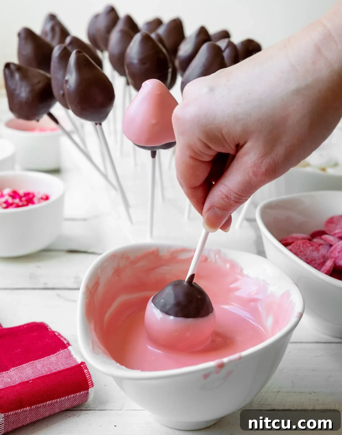 valentines-day-treat A hand carefully dips a brown chocolate-covered strawberry pop into a bowl of melted pink chocolate, creating a beautiful layered effect. This step adds vibrant color and another dimension of flavor to the treat.