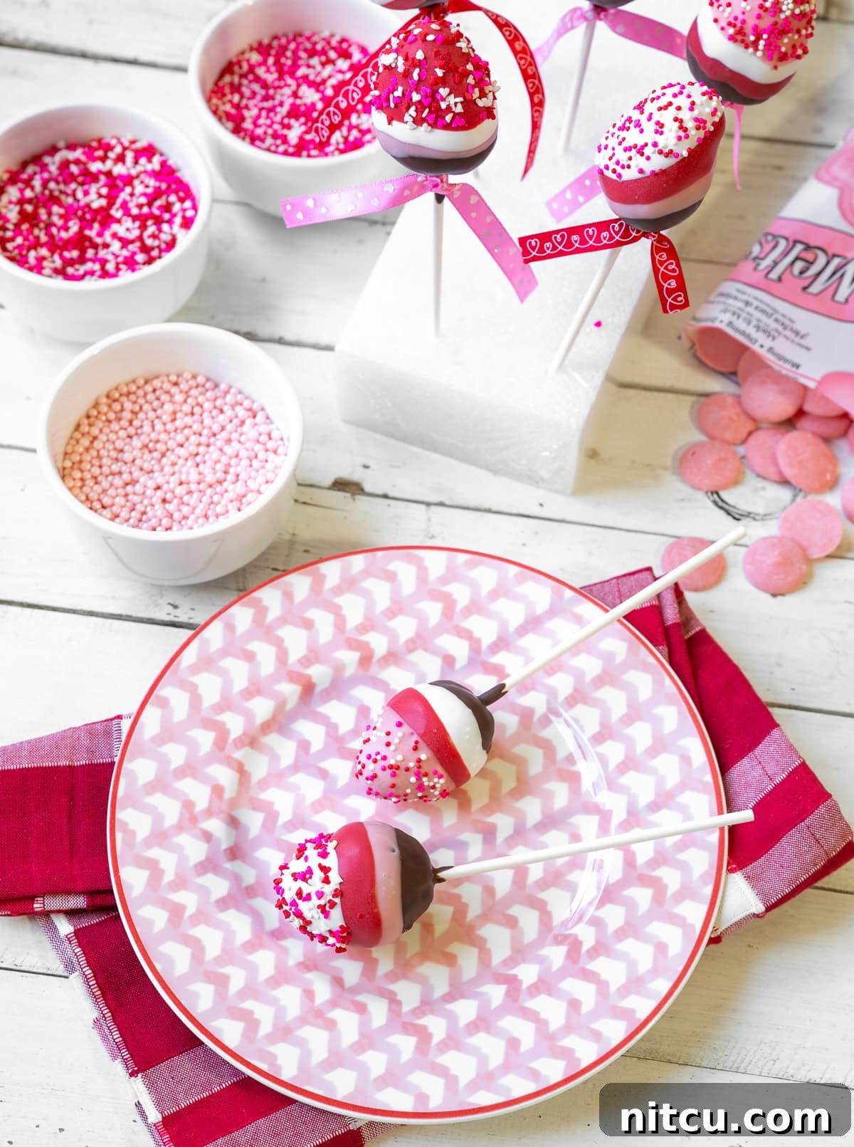 Gourmet Chocolate Strawberry Pops 5 Two exquisite chocolate-covered strawberry lollipops, beautifully layered with rich brown chocolate, delicate pink, pure white, and vibrant red, all artfully topped with charming pink and white Valentine's Day sprinkles. They are presented against a soft, romantic background.