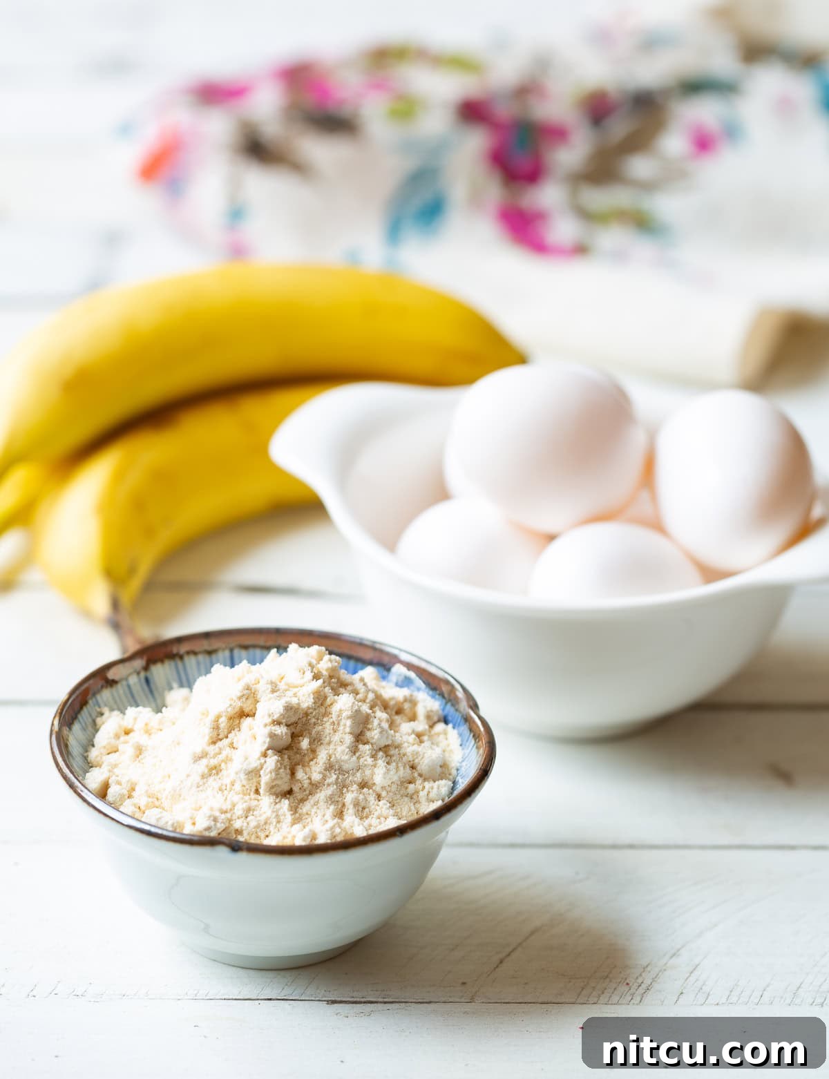 Paleo Pancake Ingredients Essential ingredients for paleo pancakes: a small bowl of white coconut flour, six fresh eggs in a bowl, and a bunch of ripe bananas, ready for blending.
