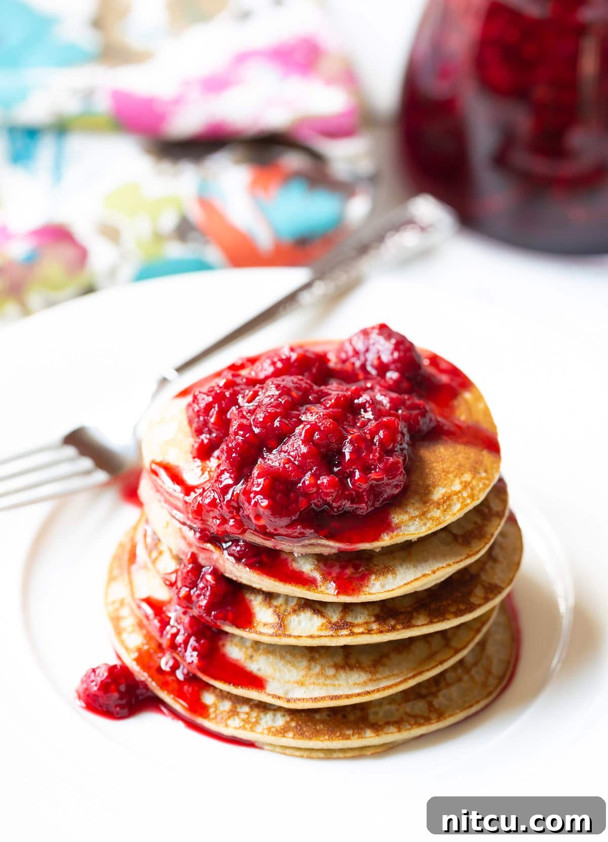 Coconut Flour Pancakes with Raspberry Syrup A stack of five perfectly cooked coconut flour pancakes, generously topped with vibrant red raspberry syrup, presented on a clean white plate.
