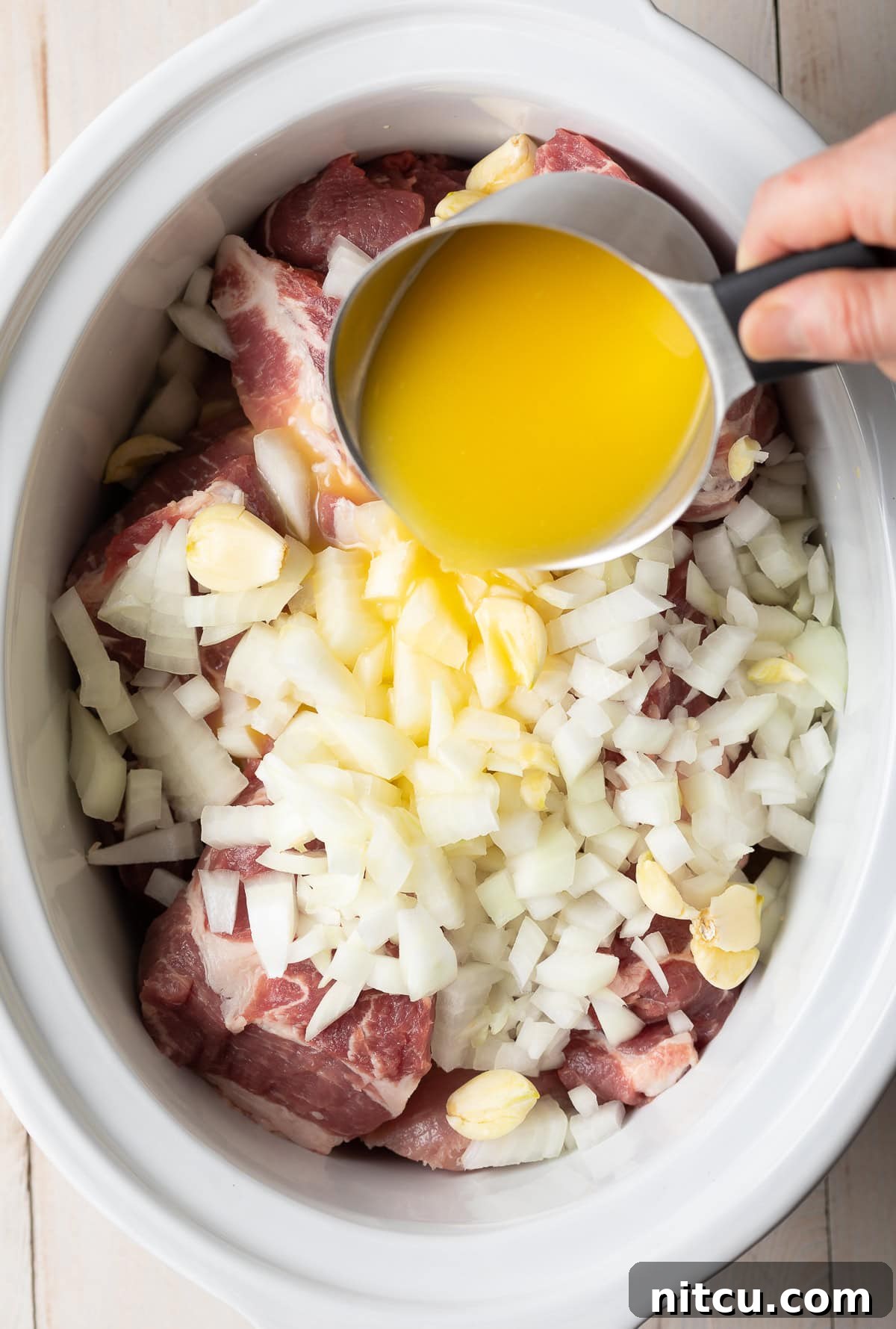 Crispy Pork Carnitas Made Easy 4 A hand pouring a cup of fresh orange juice into a slow cooker filled with pork, garlic, and onions.