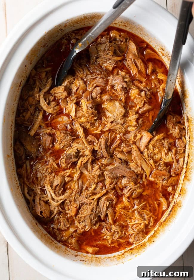 Crispy Pork Carnitas Made Easy 6 Tongs gently shredding the perfectly cooked carnitas inside the slow cooker, submerged in its flavorful juices.