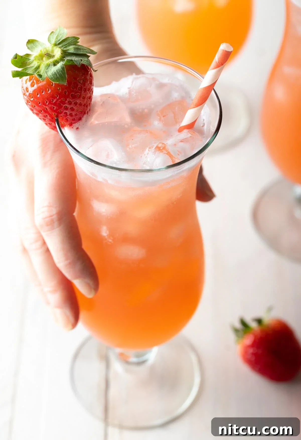 Crafting Refreshing Agua Fresca 2 Hand holding a tall cocktail glass filled with strawberry pineapple agua fresca. The glass is garnished with a strawberry on the rim and and orange and white striped paper straw.