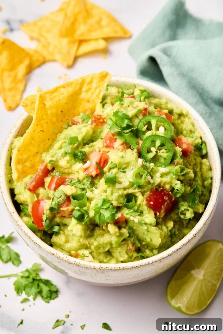 Homemade Guacamole Recipe Freshly made guacamole topped with cilantro and diced tomatoes in a white bowl, surrounded by crispy tortilla chips.