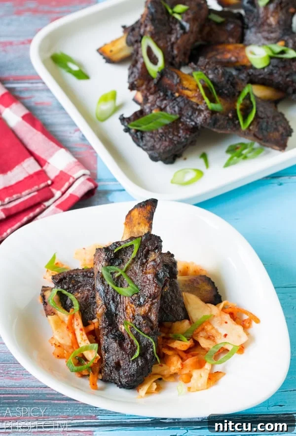 Sticky Soy-Glazed Asian Short Ribs 2 Asian beef ribs on a white plate with garnishes including kimchi and slivers of scallions.